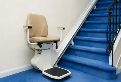 A beige stairlift chair is installed next to a staircase with bright blue carpet and a black handrail.
