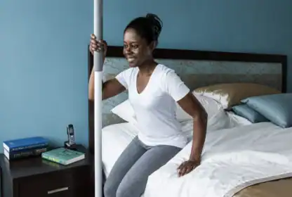 A woman sits on the edge of a bed, holding a support pole for balance. A nightstand with books and a phone is next to her.