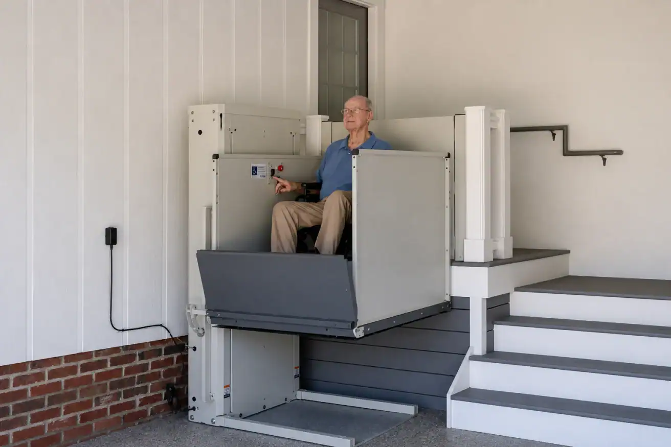 An older man in a wheelchair uses a vertical platform lift to move between ground level and a porch next to a staircase.