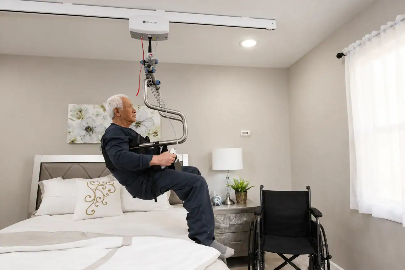 An elderly man is being lifted from a bed with a ceiling-mounted patient lift; a wheelchair is positioned beside the bed in a well-lit bedroom.