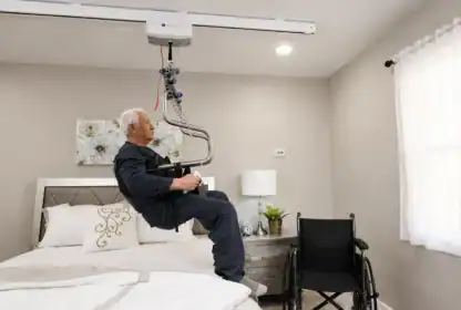 An elderly man is being lifted from a bed with a ceiling-mounted patient lift; a wheelchair is positioned beside the bed in a well-lit bedroom.