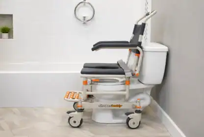 A wheeled shower commode chair labeled "shower buddy" is positioned over a toilet in a modern bathroom with white tiles and a bathtub.