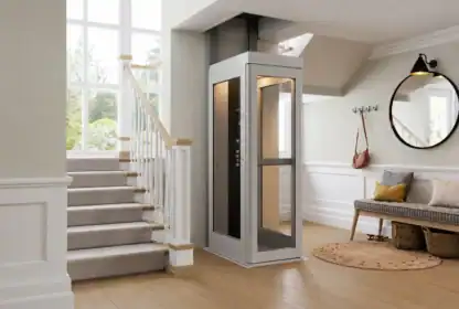 A modern home interior with a small residential elevator next to a carpeted staircase, a bench, round mirror, and large windows letting in natural light.