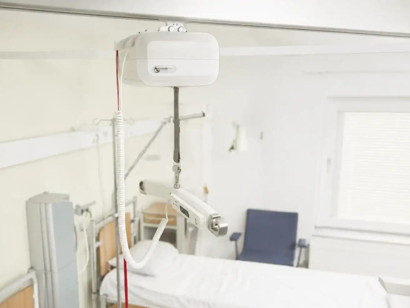 Ceiling-mounted patient lift system installed above a hospital bed in a brightly lit, unoccupied hospital room.