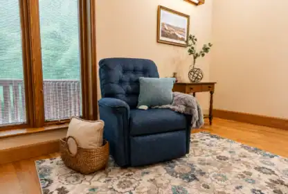 A blue recliner with pillows and a blanket sits on a patterned rug near a window; a basket with a pillow and a small table with decor are in the background.