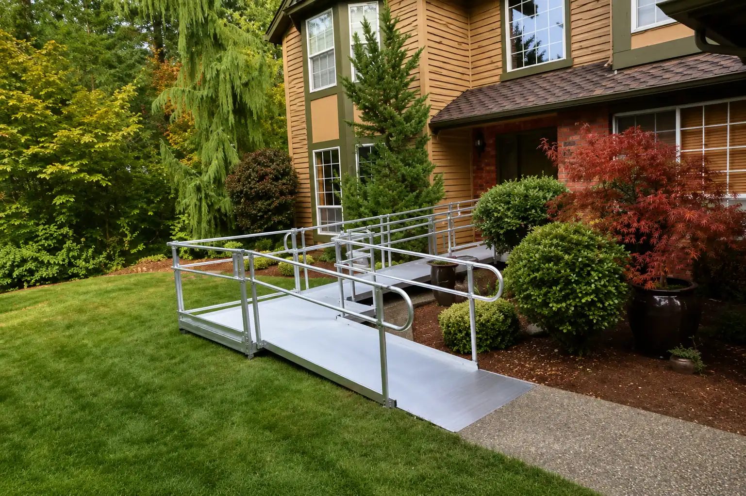 A metal wheelchair ramp with handrails leads to the entrance of a house, surrounded by green grass, bushes, and trees.