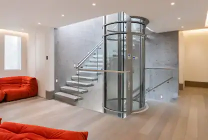 Modern interior with a circular glass elevator and floating concrete staircase, light wood flooring, and red lounge seating.