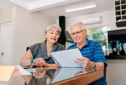 Two older adults sit at a table indoors, looking at and discussing a document together.