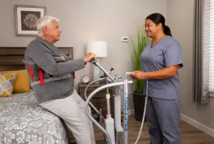 An elderly man wearing a transfer harness sits on a bed while a caregiver in scrubs operates a patient lift device beside him in a bedroom.