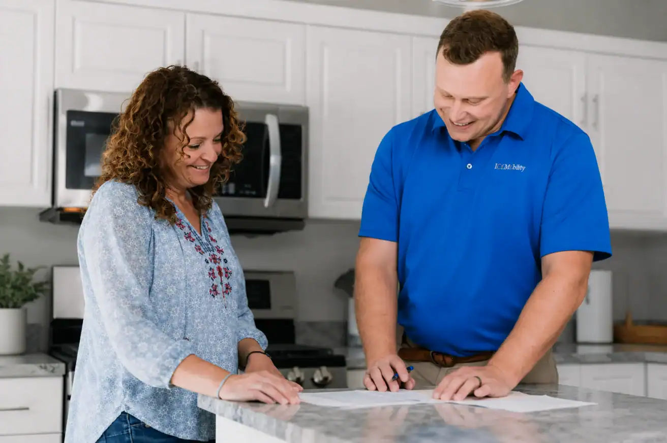 A woman and a man stand at a kitchen counter, reviewing and signing documents together. The man is wearing a blue polo shirt with a logo.
