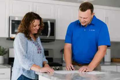 A woman and a man stand at a kitchen counter, reviewing and signing documents together. The man is wearing a blue polo shirt with a logo.