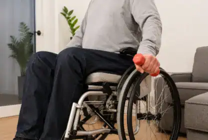 A person in a wheelchair holds a red dumbbell indoors, near a gray sofa and a potted plant.