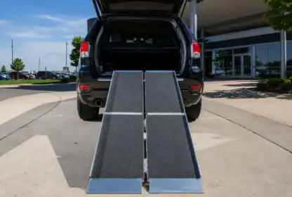 A black SUV with its rear hatch open and two metal ramps extended from the trunk to the pavement, positioned in a parking lot near a building.