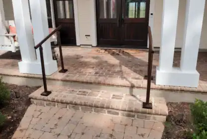 A brick front porch with two handrails on either side of three steps, leading up to double wooden doors with glass panels.