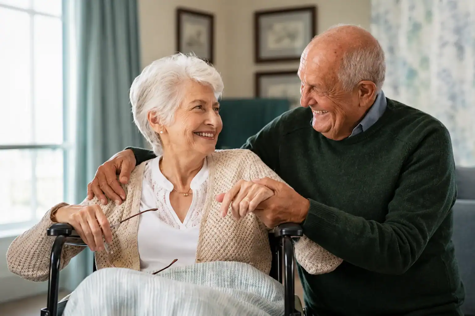 Elderly couple smiling at each other, with the woman sitting in a wheelchair and the man standing beside her, holding her hand in a cozy, well-lit room.