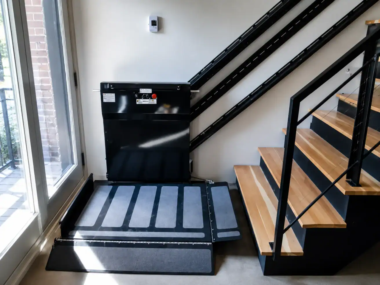 A black platform wheelchair lift installed next to a staircase, with metal rails running up the wall beside wooden steps.
