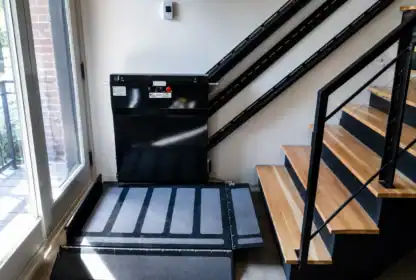 A black platform wheelchair lift installed next to a staircase, with metal rails running up the wall beside wooden steps.