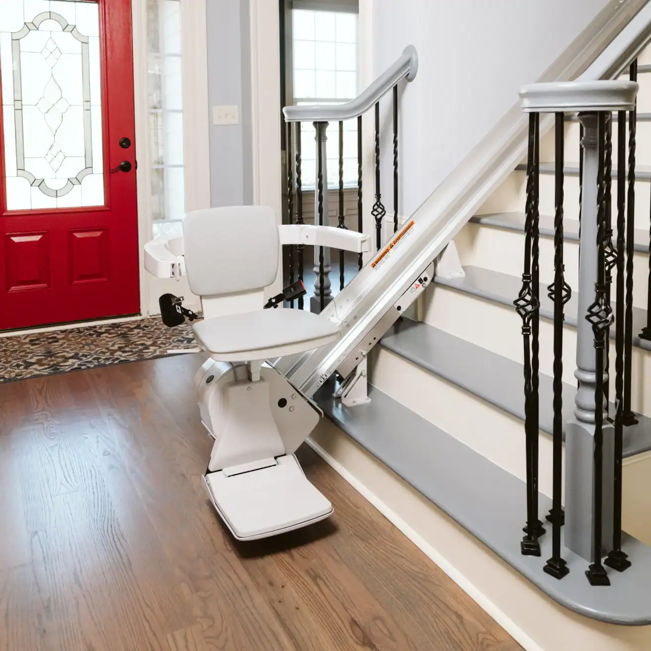 A motorized stair lift installed on a residential staircase with a red front door and wooden floor visible in the entryway.