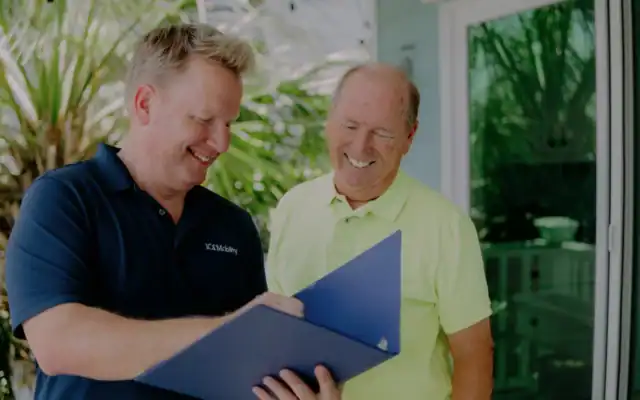 Two men standing outside, smiling and looking at documents in a blue folder. One man wears a dark polo shirt, the other a light yellow polo shirt. Green plants and a glass door are in the background.