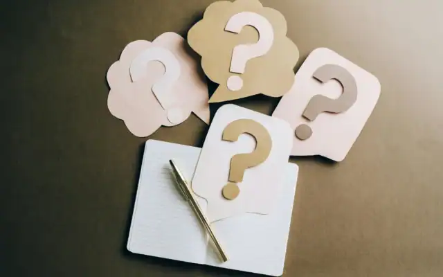 A notebook and pen are placed on a surface with several paper cutouts of speech bubbles containing question marks arranged around them.