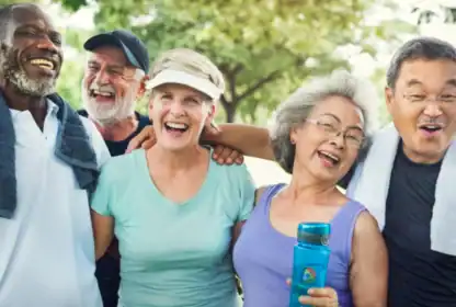 Five older adults stand close together outdoors, smiling and laughing. Two have towels around their necks, and one holds a water bottle. Trees and greenery are visible in the background.