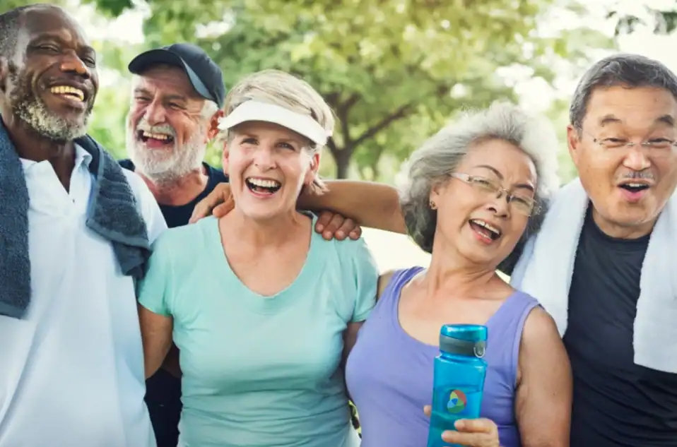 Five older adults stand close together outdoors, smiling and laughing. Two have towels around their necks, and one holds a water bottle. Trees and greenery are visible in the background.