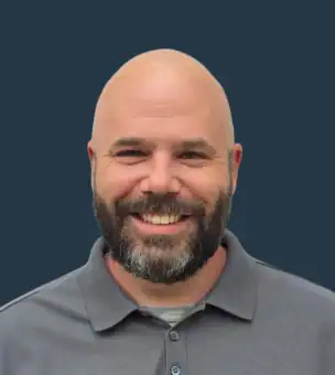 A smiling bald man with a beard wearing a gray collared shirt stands in front of a plain dark background.
