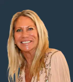 A woman with long blonde hair, wearing a light-colored embroidered blouse, smiles at the camera against a plain dark background.