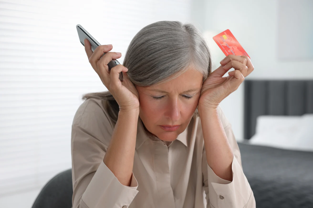 A woman with gray hair holds a phone in one hand and a credit card in the other, looking stressed with her eyes closed.