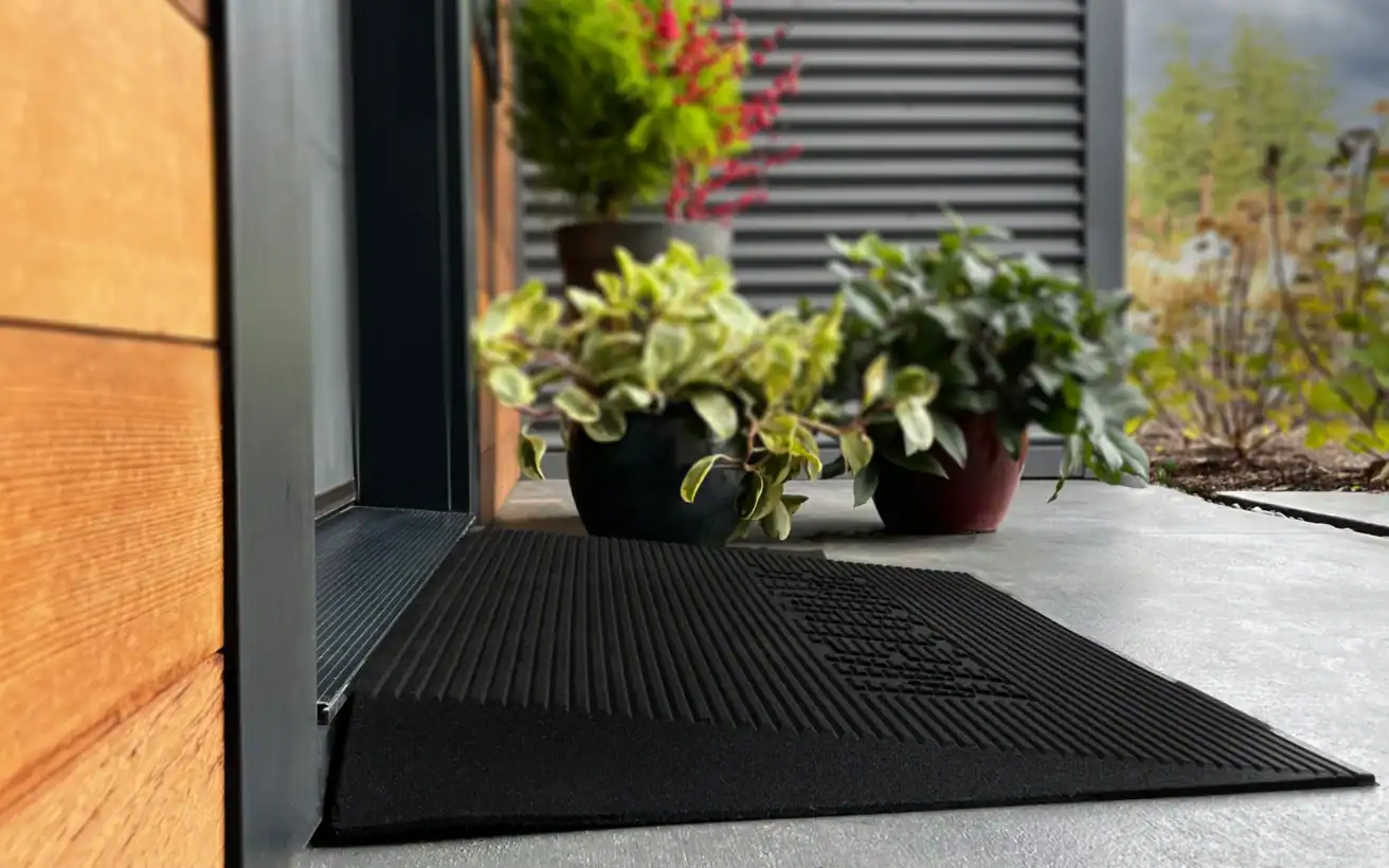 A black doormat sits at the entrance of a modern home, with potted plants arranged on the concrete porch next to the door.
