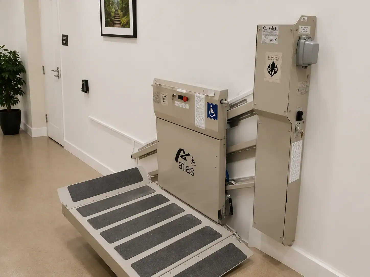 Wall-mounted wheelchair lift with a folded ramp, control panel, and safety instructions in a hallway with beige flooring.