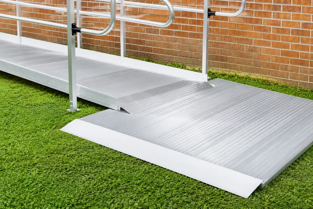 A metal wheelchair ramp with handrails installed over grass, leading up to a brick wall building.