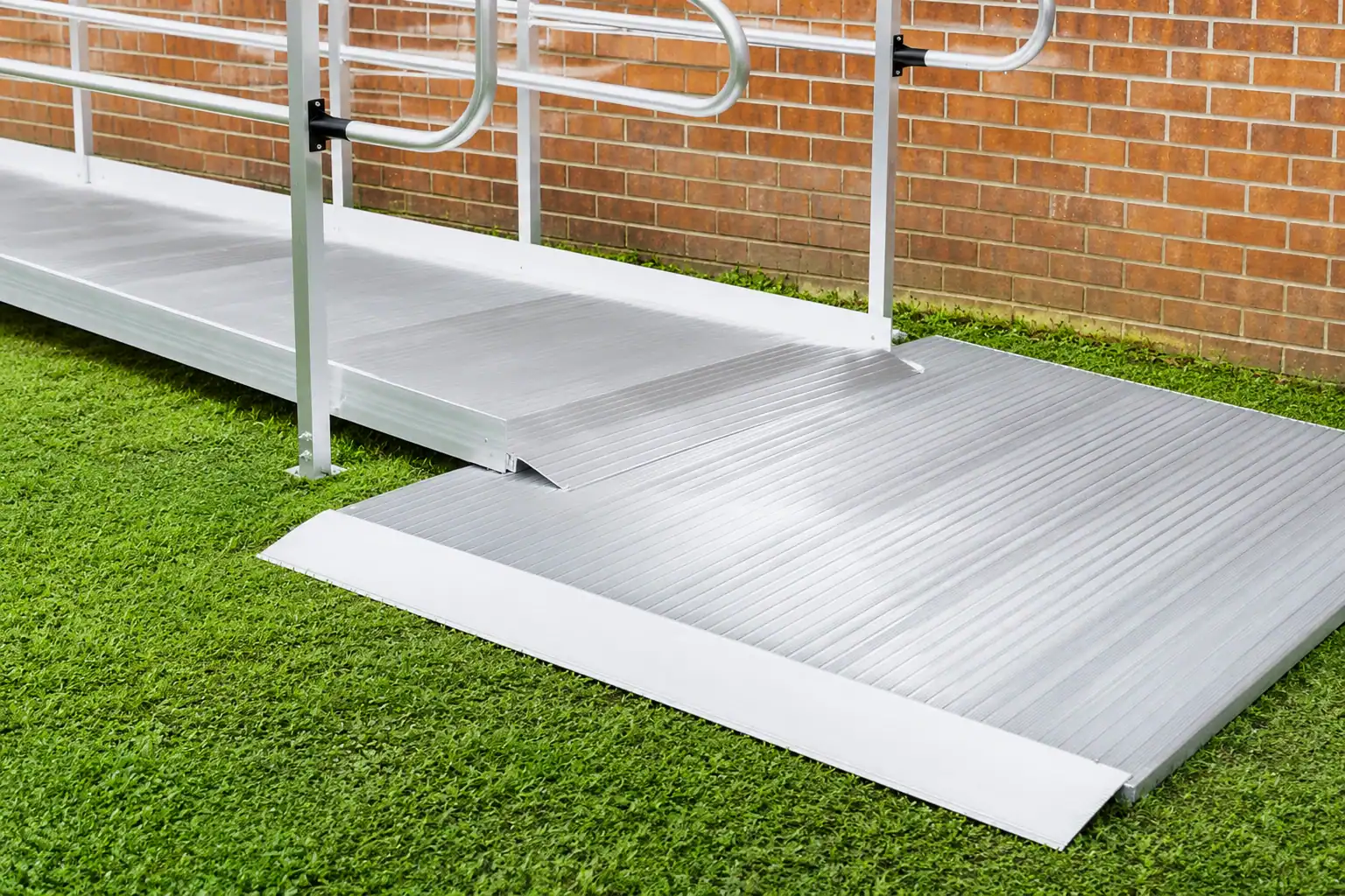 A metal wheelchair ramp with handrails installed over grass, leading up to a brick wall building.