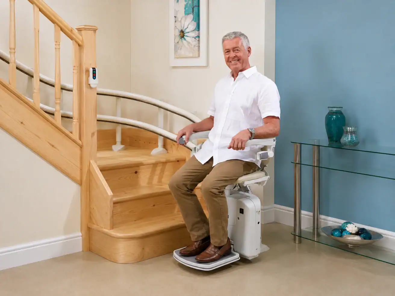Older man in a white shirt and khaki pants sits on a motorized stairlift at the base of a wooden staircase in a modern home setting.