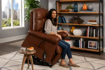 A woman sits in a brown leather recliner with a remote control in a living room, next to a small table with a glass of water and fruit, and a bookshelf in the background.