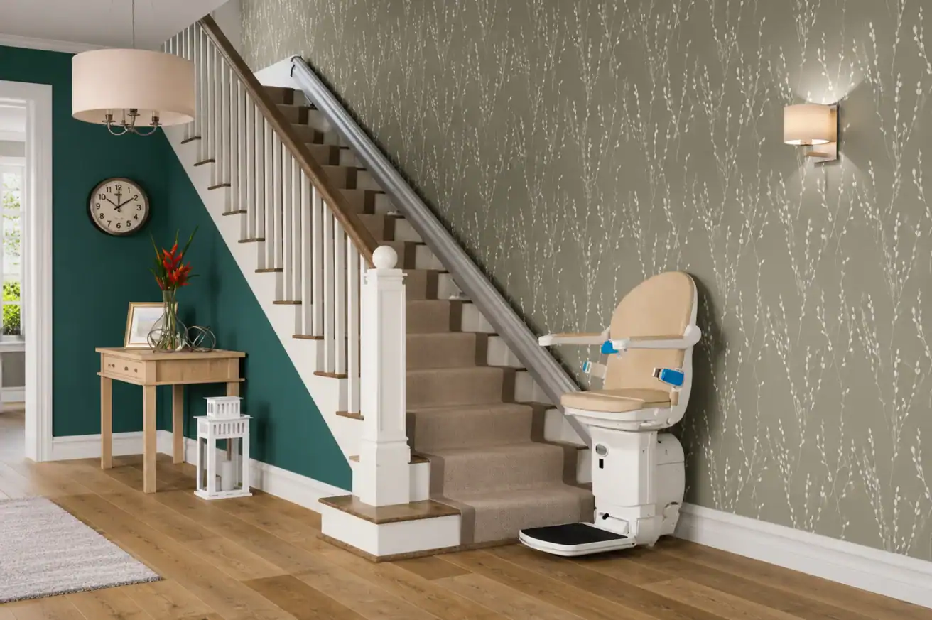 A beige stairlift is installed along a carpeted staircase in a modern home with green and patterned wallpaper, wooden flooring, and decorative furnishings.