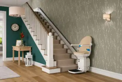 A beige stairlift is installed along a carpeted staircase in a modern home with green and patterned wallpaper, wooden flooring, and decorative furnishings.
