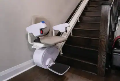 A stairlift with a padded seat and armrests is installed beside a dark wooden staircase in a residential setting.