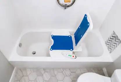 A bathtub with a blue and white electric bath lift chair inside, surrounded by white tile walls and hexagonal floor tiles.