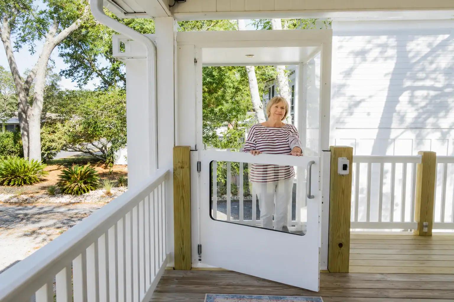 A woman stands on a porch, holding open a white gate style door attached to the entrance, with trees and a white house visible in the background.