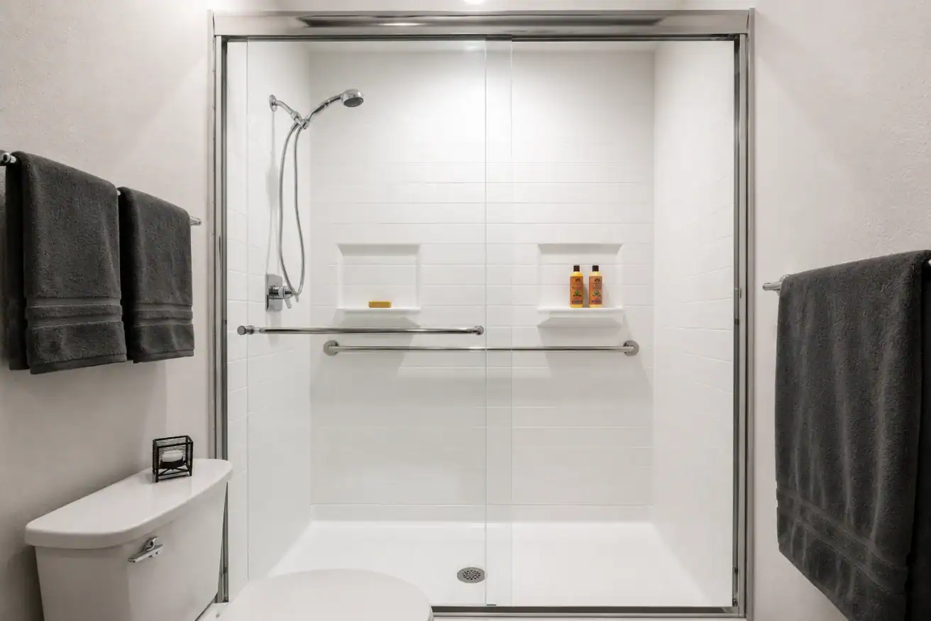 A modern bathroom with a glass-enclosed shower, two wall shelves holding toiletries, a mounted handheld showerhead, metal grab bars, and dark towels on towel racks.