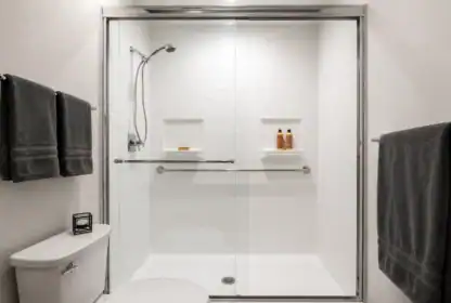 A modern bathroom with a glass-enclosed shower, two wall shelves holding toiletries, a mounted handheld showerhead, metal grab bars, and dark towels on towel racks.