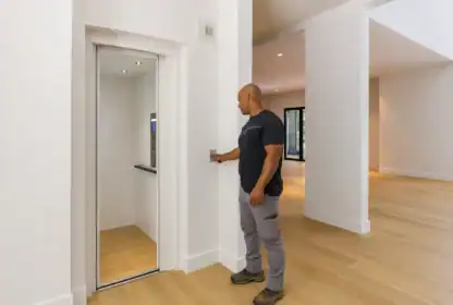 A man stands in a modern, light-filled room pressing the button to operate a small residential elevator with open doors.