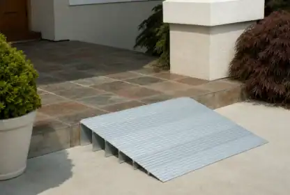 A metal wheelchair ramp is positioned over a step leading to a tiled porch, with potted plants on either side.
