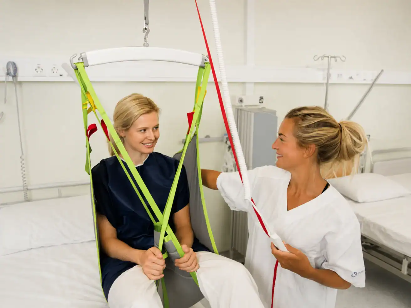 Two healthcare workers in a hospital room demonstrate a patient lifting sling, with one sitting in the sling and the other operating the equipment.