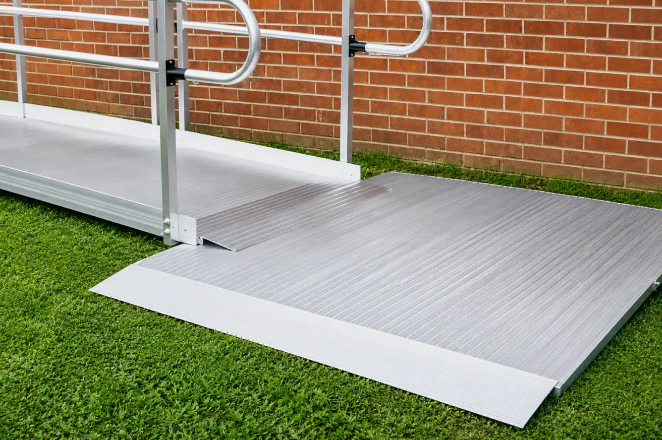 A metal wheelchair ramp with handrails is installed over grass, leading up to a brick wall.