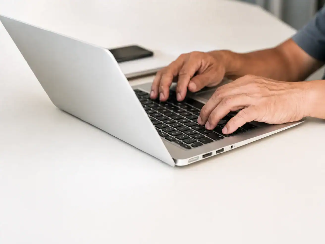 A person types on a silver laptop at a white desk, with a smartphone placed nearby.