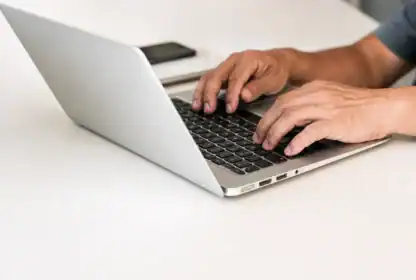 A person types on a silver laptop at a white desk, with a smartphone placed nearby.