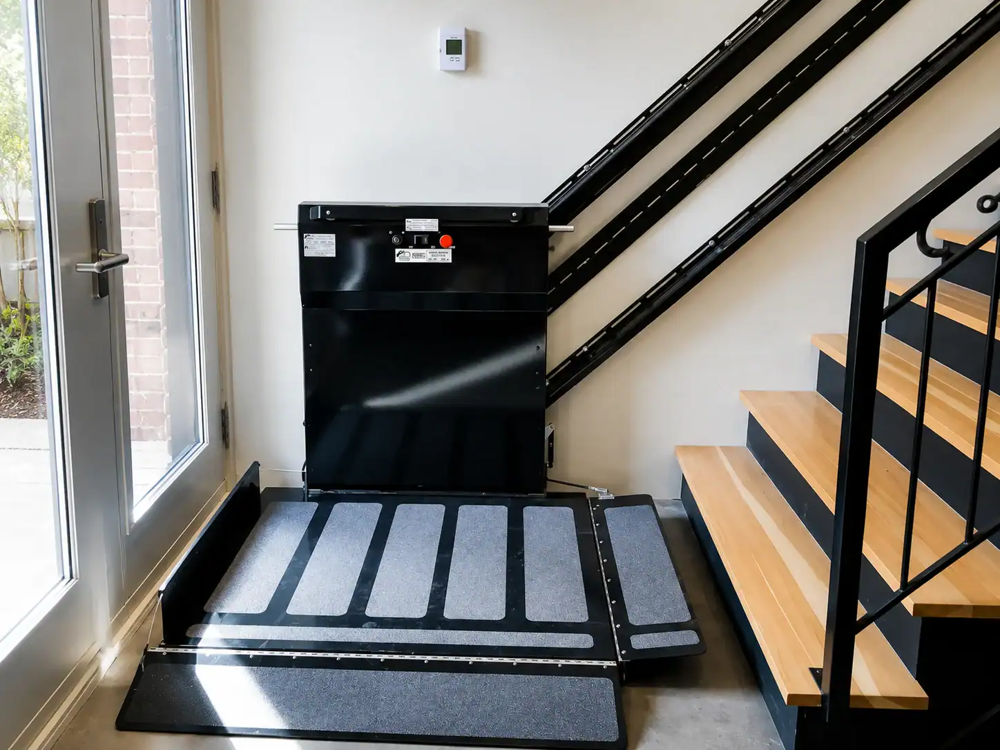 A wheelchair lift with a ramp installed at the base of an indoor staircase, next to a glass door and railings.