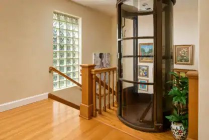 A modern glass and metal home elevator is installed beside a wooden staircase in a well-lit hallway with framed artwork and a potted plant.